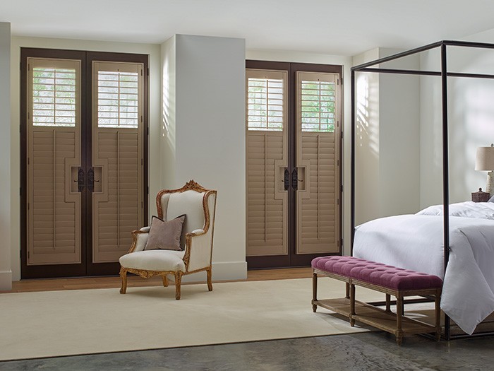 Large room with brown wooden shutters opened one quarter of the way in front on french doors in front of bed with black railing and white comforter. 